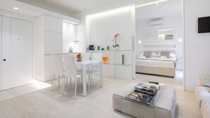 Un espacio habitable moderno y minimalista con comedor, una mesa de centro de vidrio y un dormitorio con cama y mesita de noche.