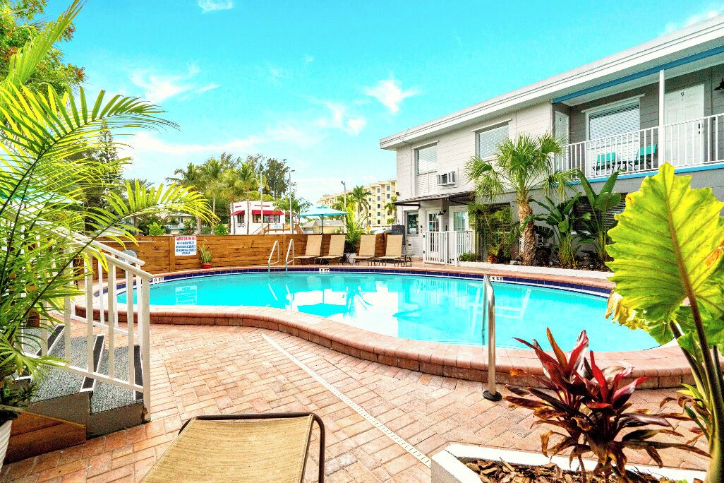A swimming pool with a brick deck, surrounded by plants and a building with a balcony.