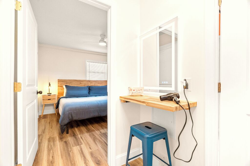 A bedroom with a bed, bedside table, lamp, and a desk with a mirror and hairdryer.