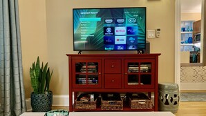 A living room with a flat-screen TV on a stand, a red entertainment center with books and decorative items, a glass ashtray, and a remote control on a white table.