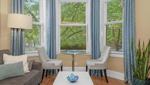 A room with a glass-top table, two chairs, a sofa, and a view of greenery through large windows.