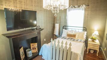 A bedroom with a television on a stand, a chandelier, a bed with a white picket fence, a bedside table with a lamp, and a window with curtains.