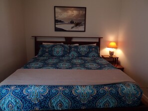 Ein Bett mit gemustertem Überwurf, einem Kopfteil, einer Nachttischlampe und einem gerahmten Bild an der Wand.
