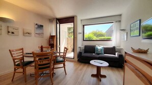 Ein gemütliches Wohnzimmer mit Essbereich, einer Couch und einem kleinen Tisch. An den Wänden hängen Bilderrahmen und ein Fenster bietet Blick ins Grüne.