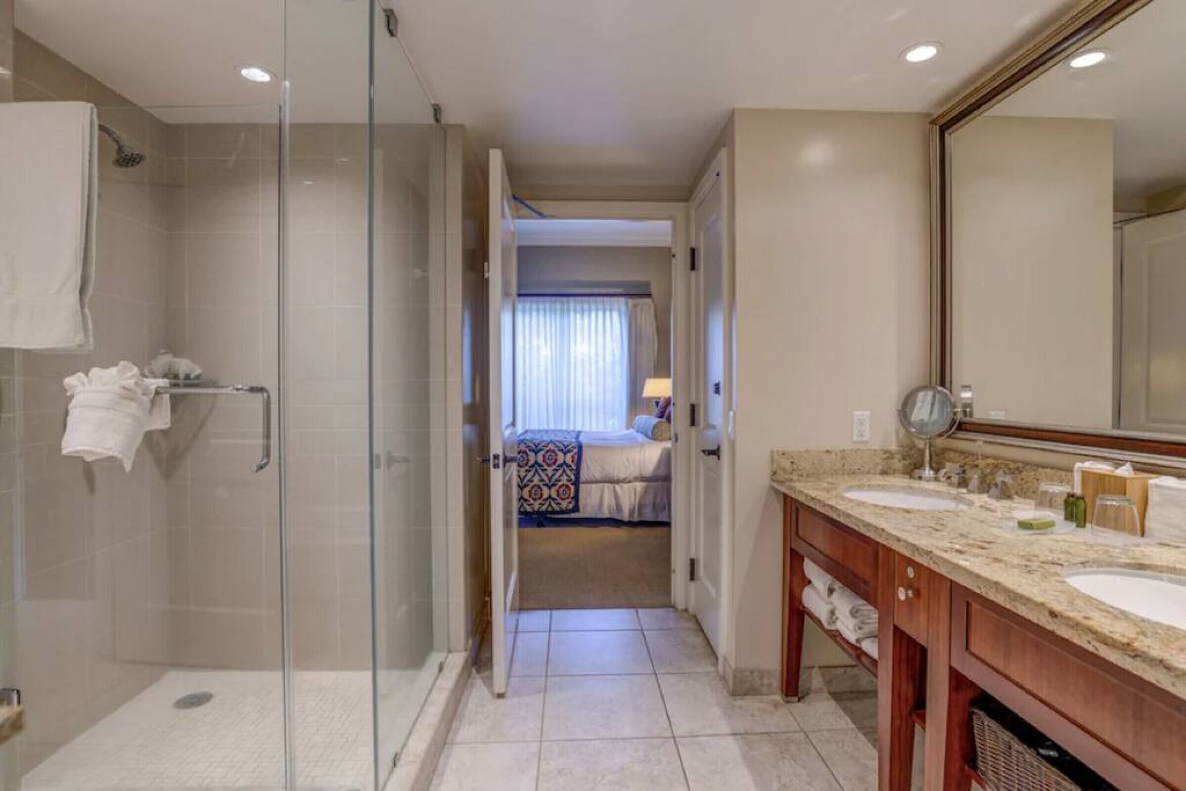 A hotel bathroom with a glass shower enclosure, a double sink vanity, and a large mirror.