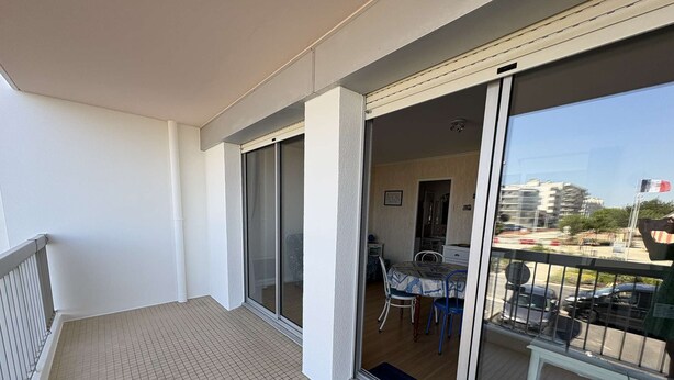 Un balcón con piso de baldosas, una puerta corrediza de vidrio que da a un área interior de comedor con mesa y sillas.