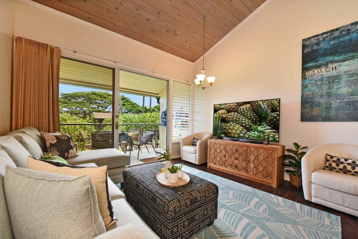 Een moderne woonkamer met een bank, poef en salontafel met dienblad. Er is een televisie aan de muur en een groot raam met zonwering.