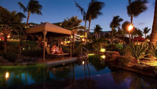 A nighttime resort pool area with palm trees, a covered seating area, and a well-lit pool.