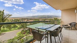 A balcony with a glass table and chairs, overlooking a green landscape with palm trees and a distant mountain range.