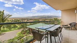 Uma varanda com mesa e cadeiras de vidro, com vista para uma paisagem verde com palmeiras e uma cordilheira ao longe.