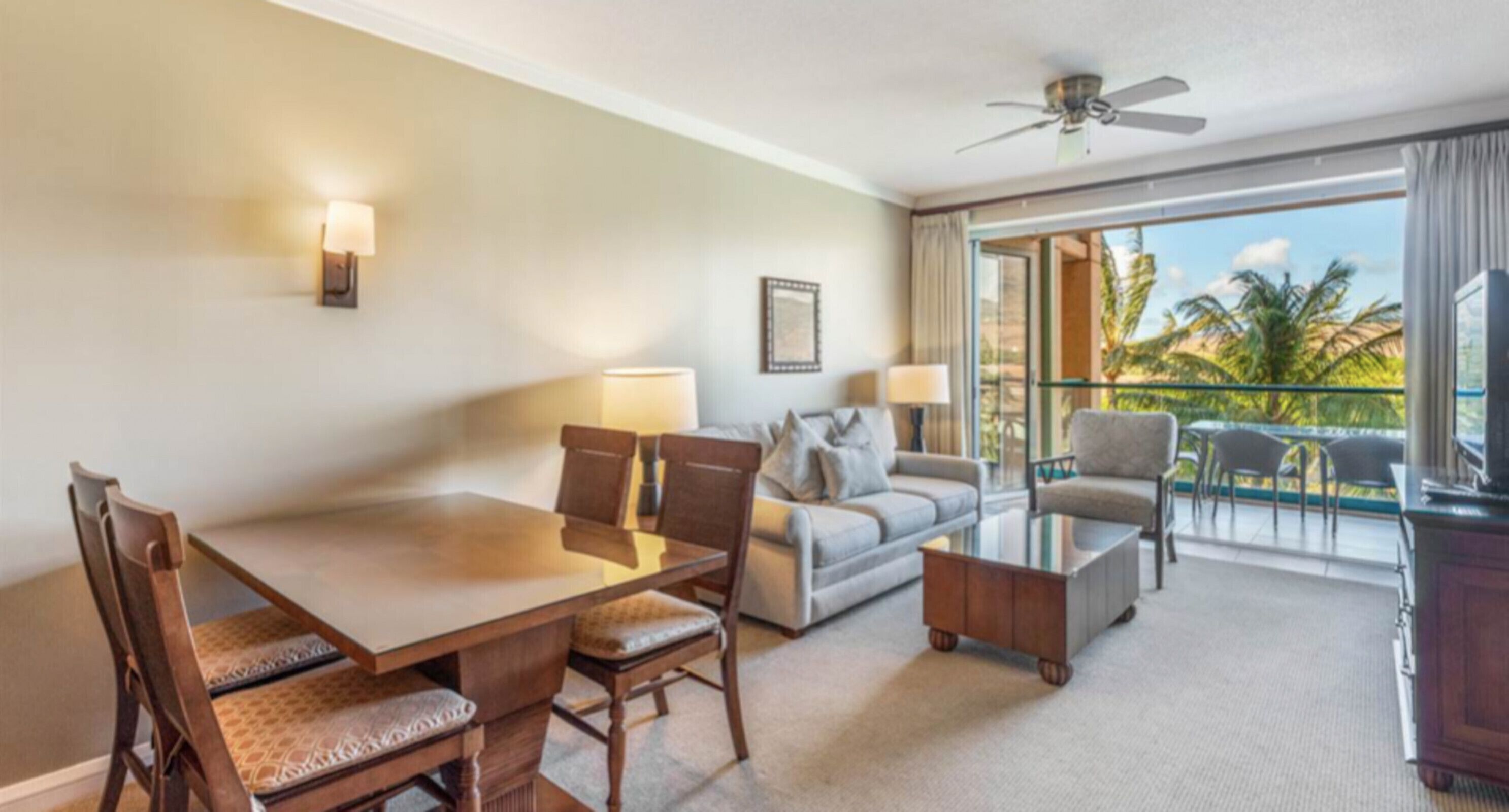 Quarto de hotel com mesa de jantar, duas cadeiras, sofá, mesa de centro e varanda com vista para palmeiras.