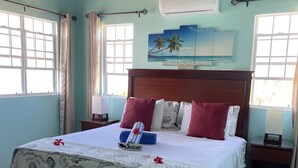 A bedroom with a large bed, a wooden ceiling, a beach-themed painting, and a window with curtains.