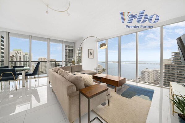 A spacious living room with a large window offering a city view, a sofa, a coffee table, and a dining area with chairs. - ICON W BRICKELL OCEAN VIEW 2BR/2BA #3310 (Miami)