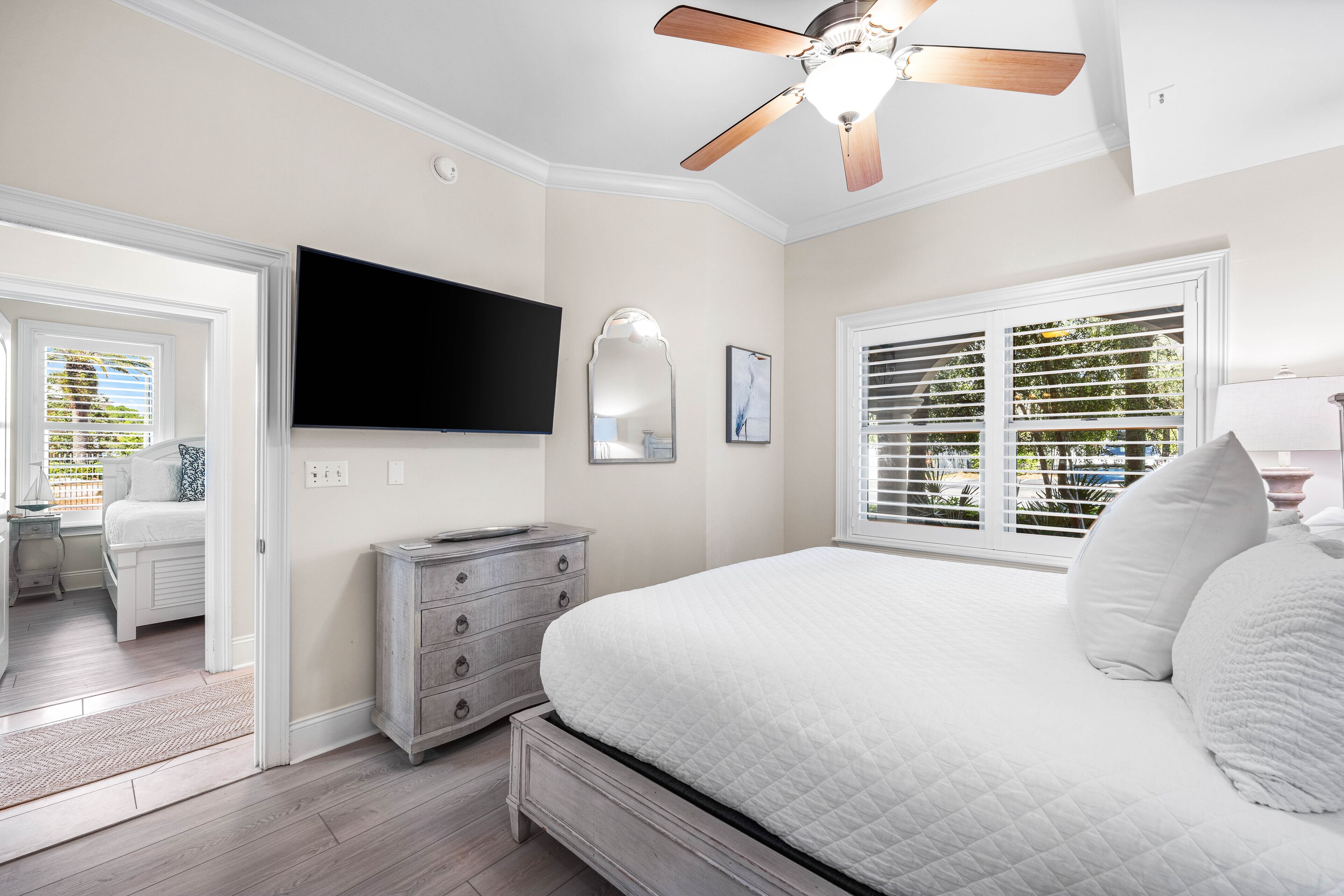 Un dormitorio con cama, cómoda, ventilador de techo, televisor y ventana con persianas.