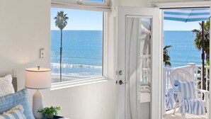 A bedroom with a view of the ocean, a door leading to a balcony, a bed with blue-striped pillows, and a lamp on a bedside table. - #1 Location, Spacious Beach House, Steps to Fun (Oceanside)