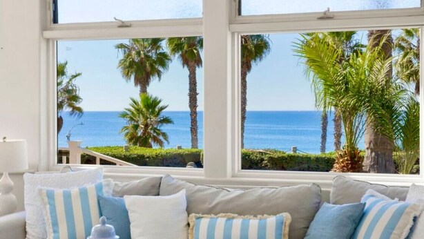 A living room with a view of the ocean, featuring a beige sofa with striped pillows and a blue vase. - #1 Location, Spacious Beach House, Steps to Fun (Oceanside)