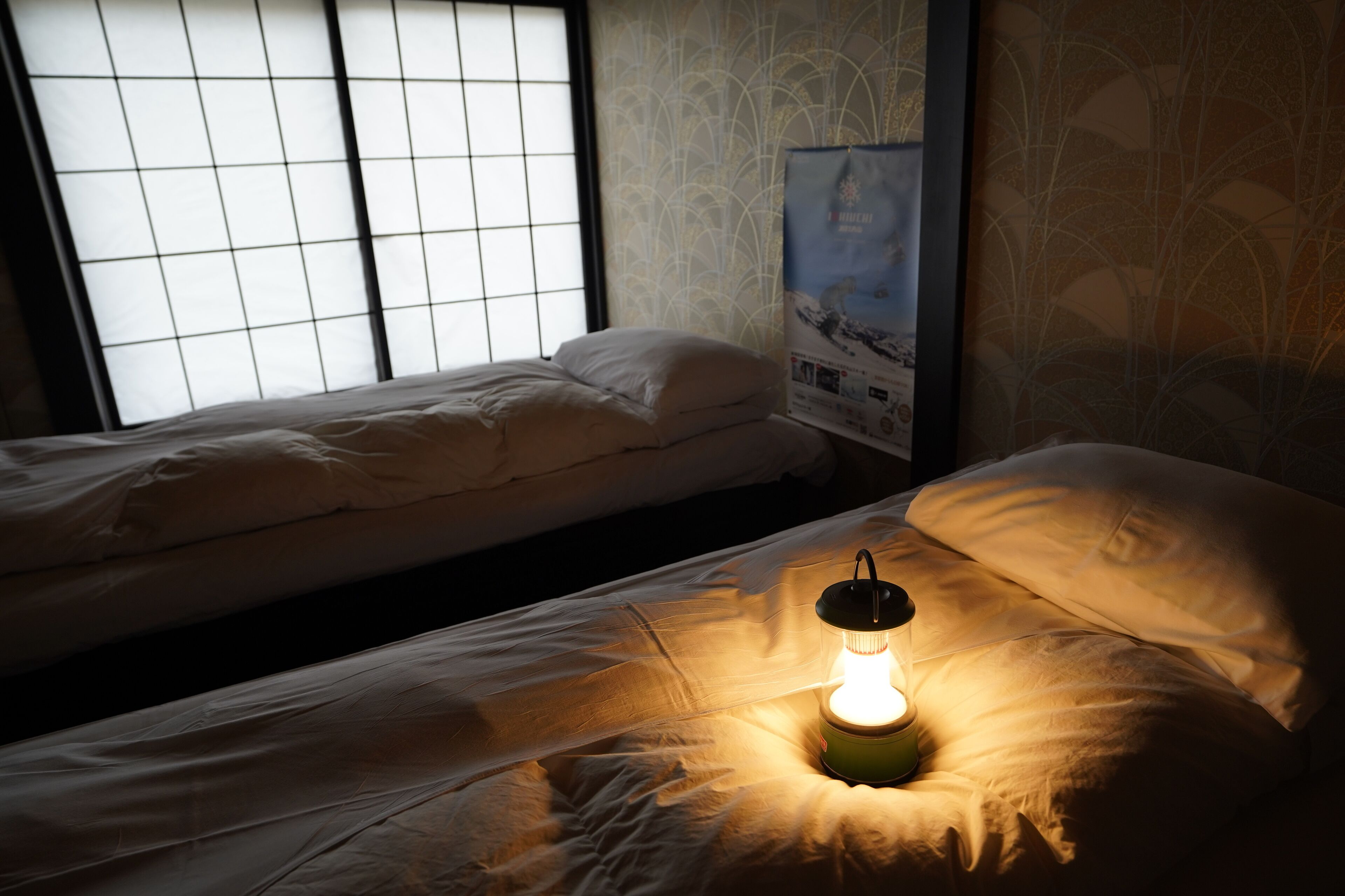 A dimly lit room with two single beds, a small table with a lamp, and a poster on the wall.