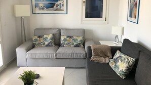 A modern living room with a grey sofa set, a white coffee table, and a potted plant. There are framed pictures on the walls and a lamp on a side table. - sam29 3-Bed Villa in Monolithos (Monolithos)