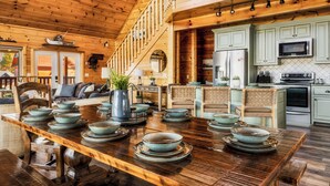 A wooden interior with a dining table set with bowls and plates, a kitchen with light green cabinets, and a staircase leading to an upper level. - Read Our Reviews! Indoor Pool + Outdoor Pool + View (Sevierville)