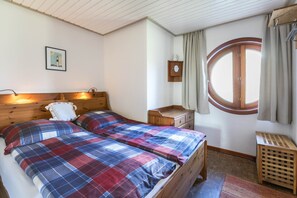 A bedroom with two beds, a wooden dresser, a small table, a window with curtains, and a wall-mounted clock.