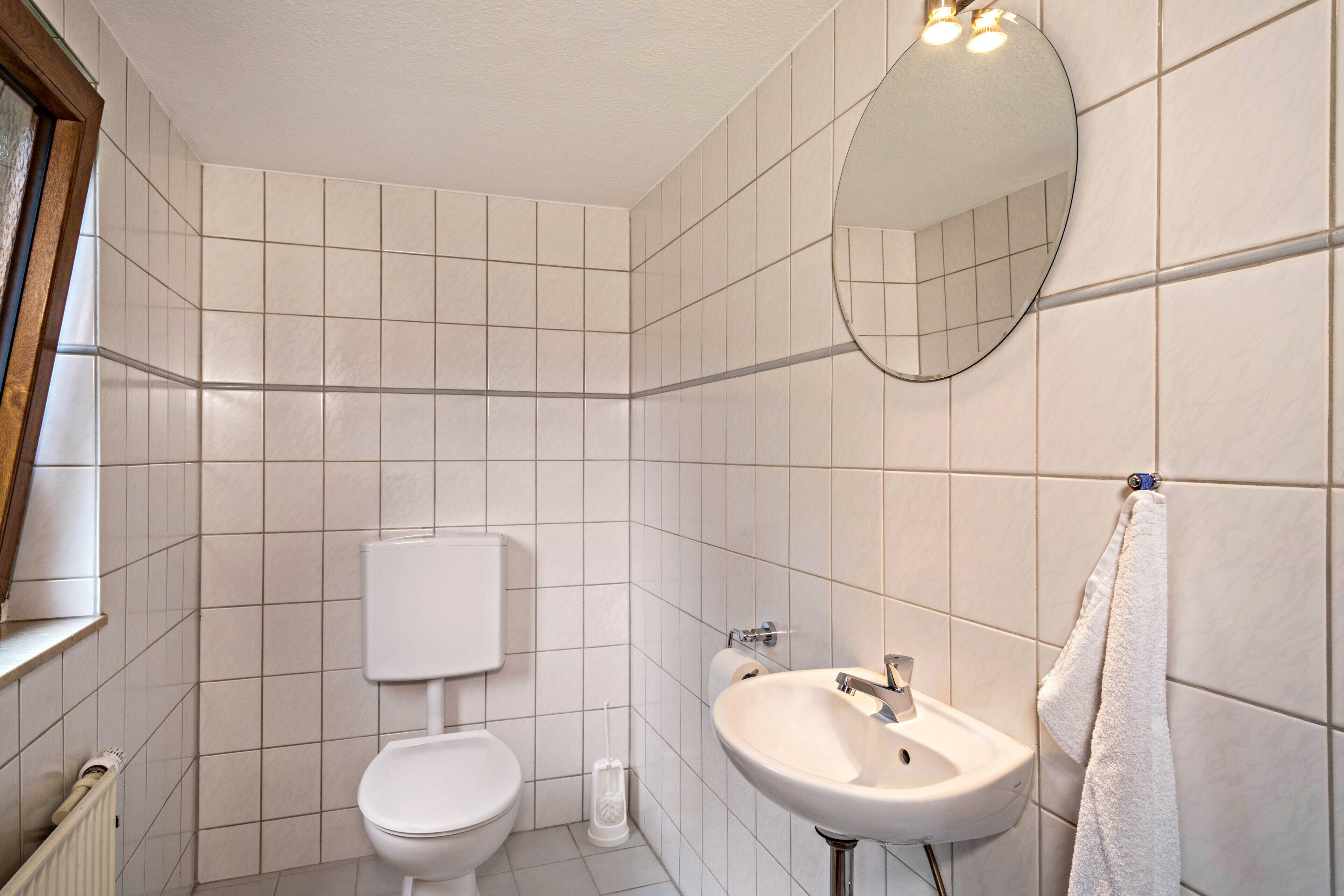 A bathroom with white tiles, a toilet, a sink, a mirror, and a towel hanging on the wall.