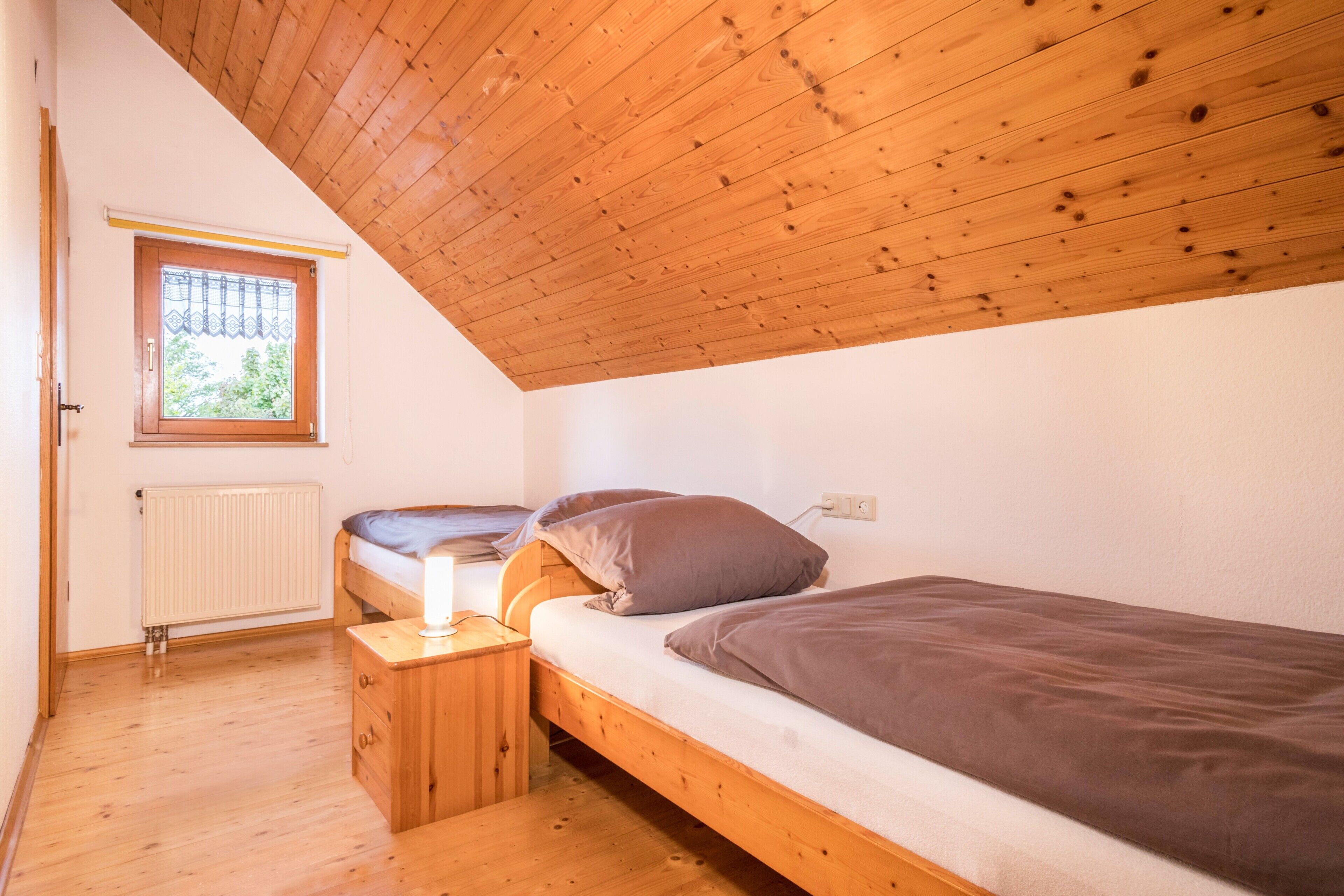 A small, single-occupancy room with a sloped ceiling, a window with a curtain, a radiator, and a bedside table with a lamp.