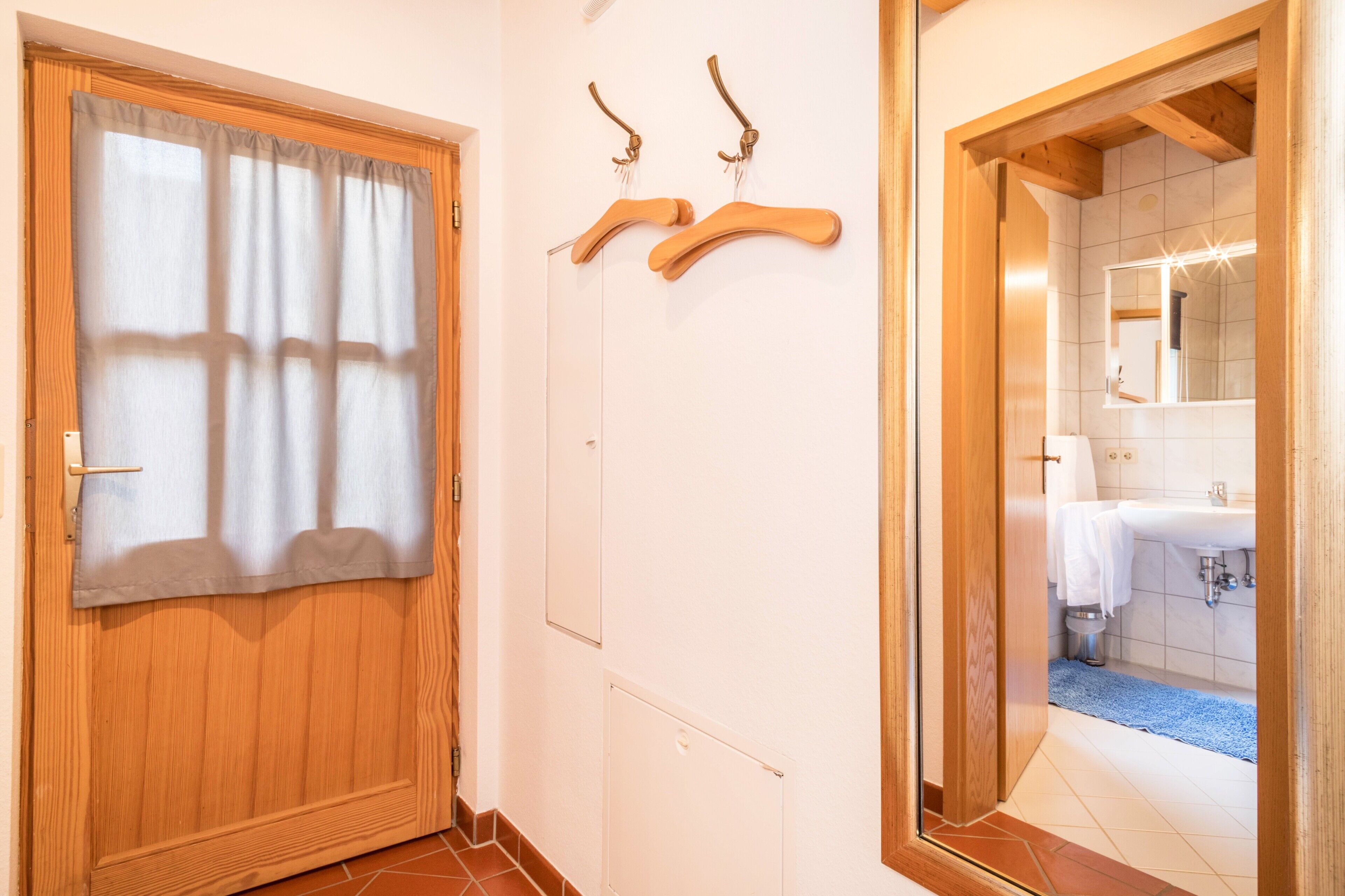 Ein Flur mit einer Holztür, einem an der Wand befestigten Kleiderhaken und einem durchgehenden Türrahmen einsehbaren Badezimmer.