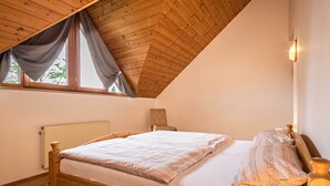 Une chambre avec un plafond en bois, un lit avec un matelas blanc et une couverture à motifs, une table de chevet avec une lampe, et une fenêtre avec des rideaux.