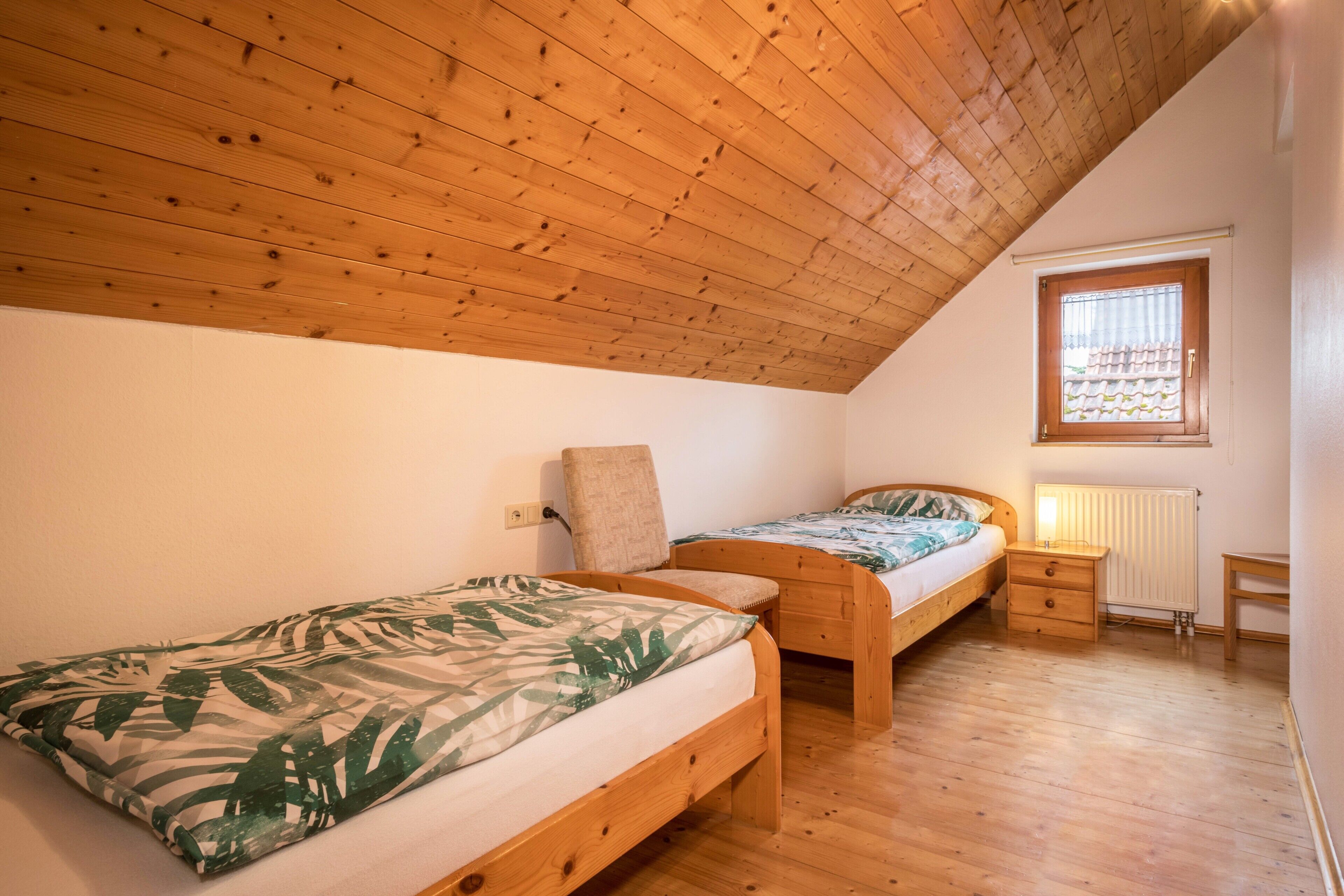 A bedroom with two wooden beds, a window with blinds, a radiator, and a wooden ceiling.