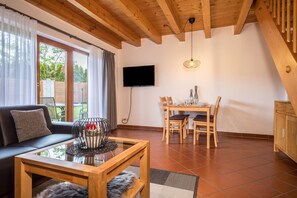 A living room with a sofa, a coffee table, a dining table with chairs, a TV mounted on the wall, and a staircase.