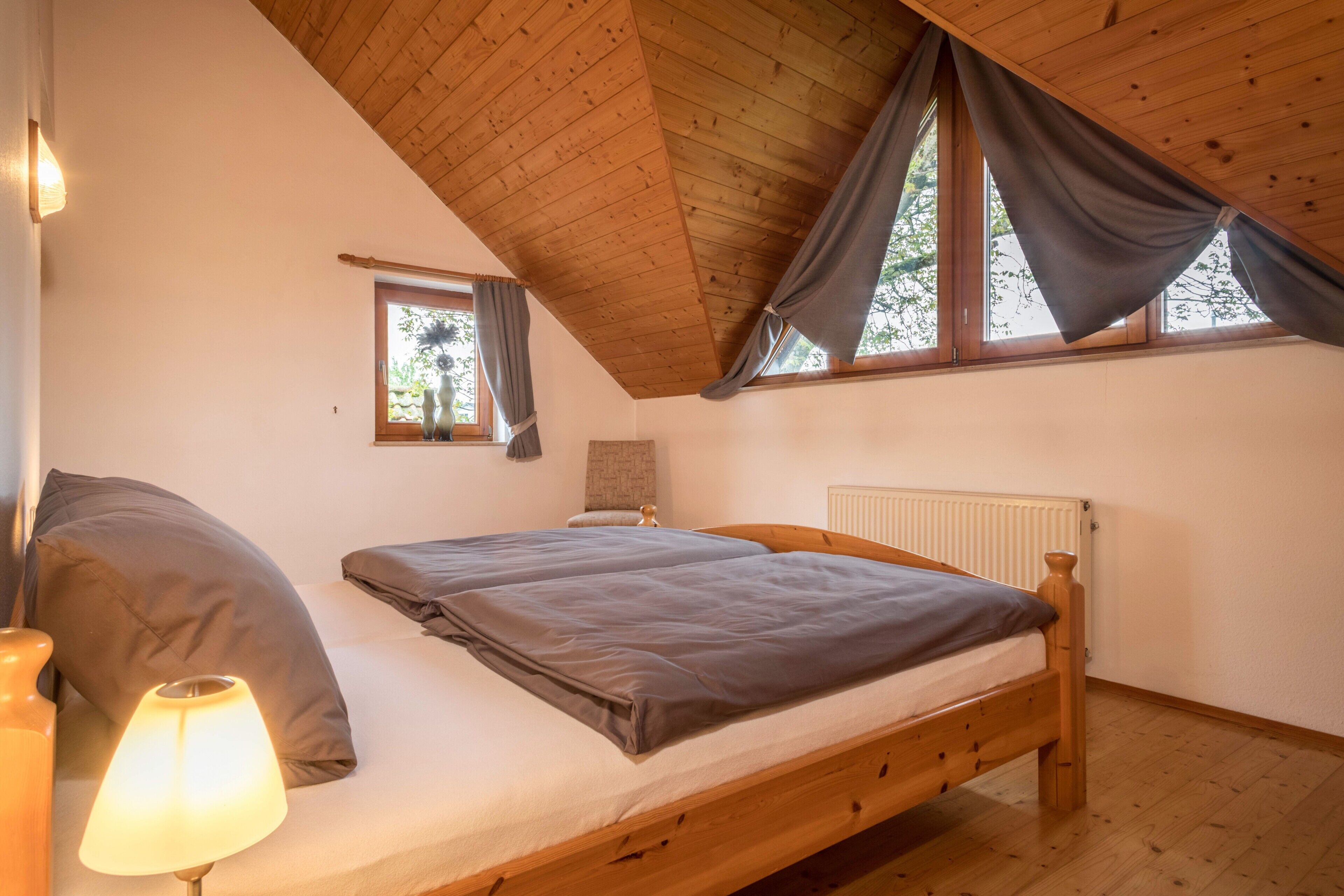 Un dormitorio con una cama de madera, una mesita de noche con una lámpara, una ventana con cortinas y un radiador.