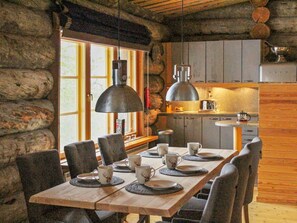 A rustic wooden dining table set with plates and cups, surrounded by chairs, in a cabin-style kitchen with stone walls and wooden beams.