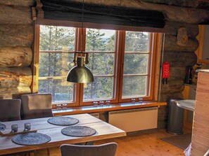 Ein rustikales Blockhäuschen im Inneren mit einem Holztisch, Stühlen, einem Fenster mit Blick auf Bäume und einer Hängelampe.