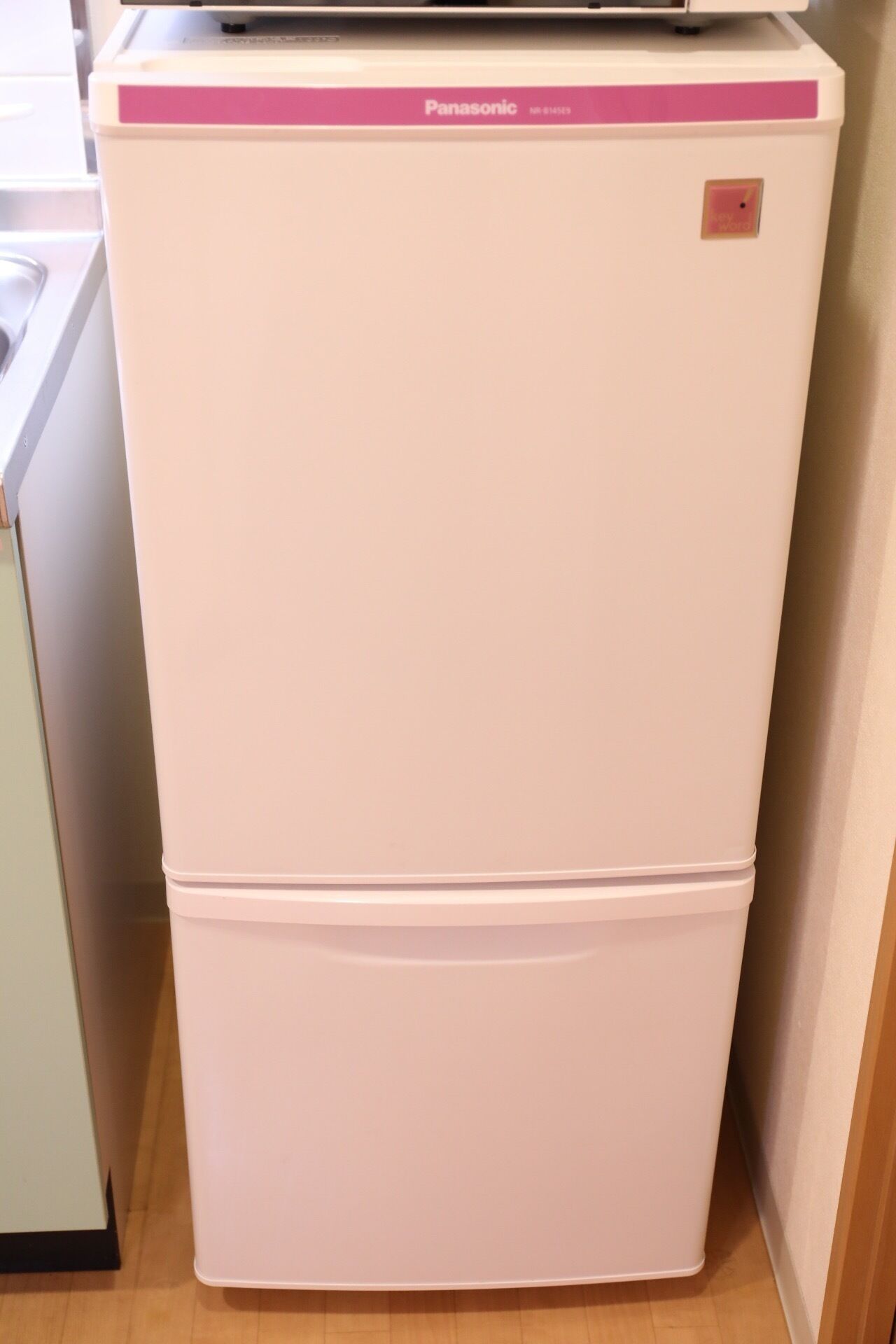 A white Panasonic refrigerator with a pink top panel, located in a kitchen with wooden flooring.