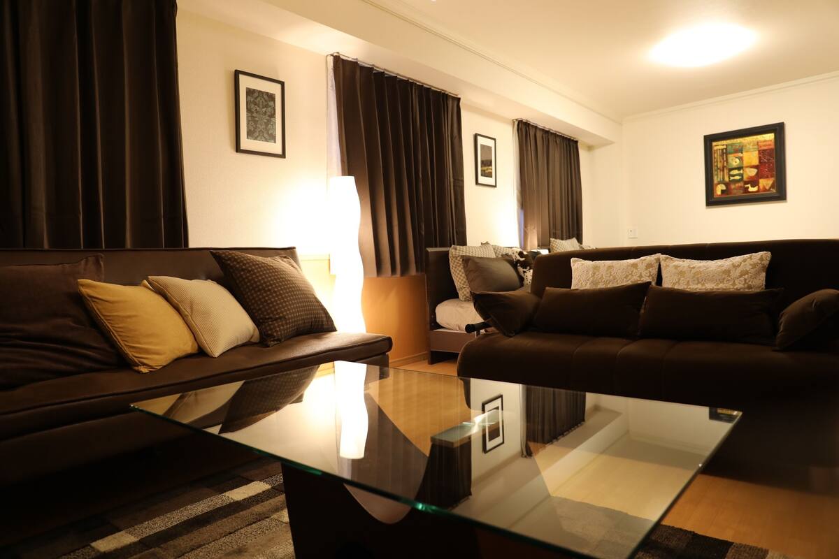 Een moderne woonkamer met een glazen salontafel, donkere banken en een lamp.