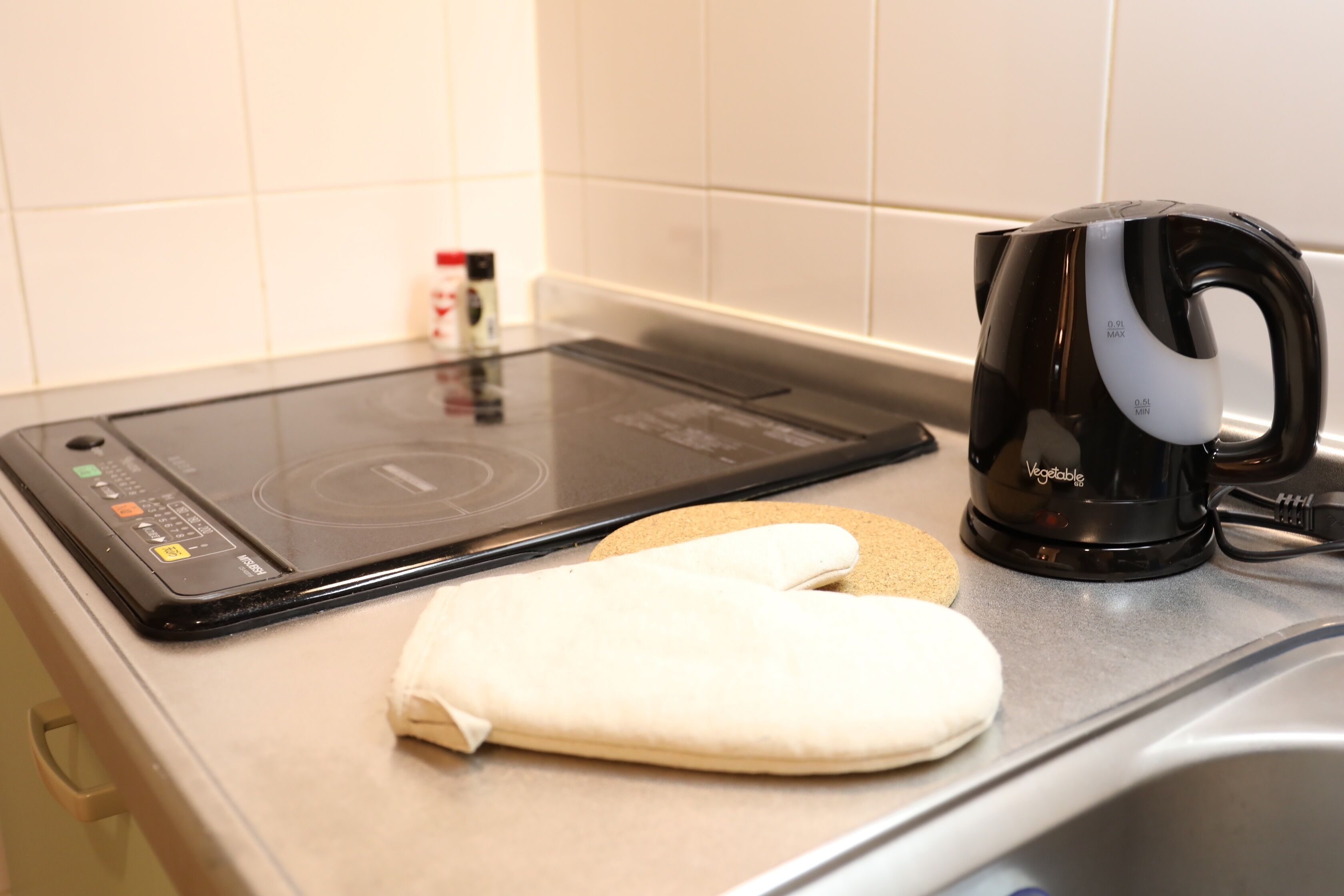 Una cocina con una placa de inducción, un hervidor negro y un guante de cocina blanco sobre la encimera.
