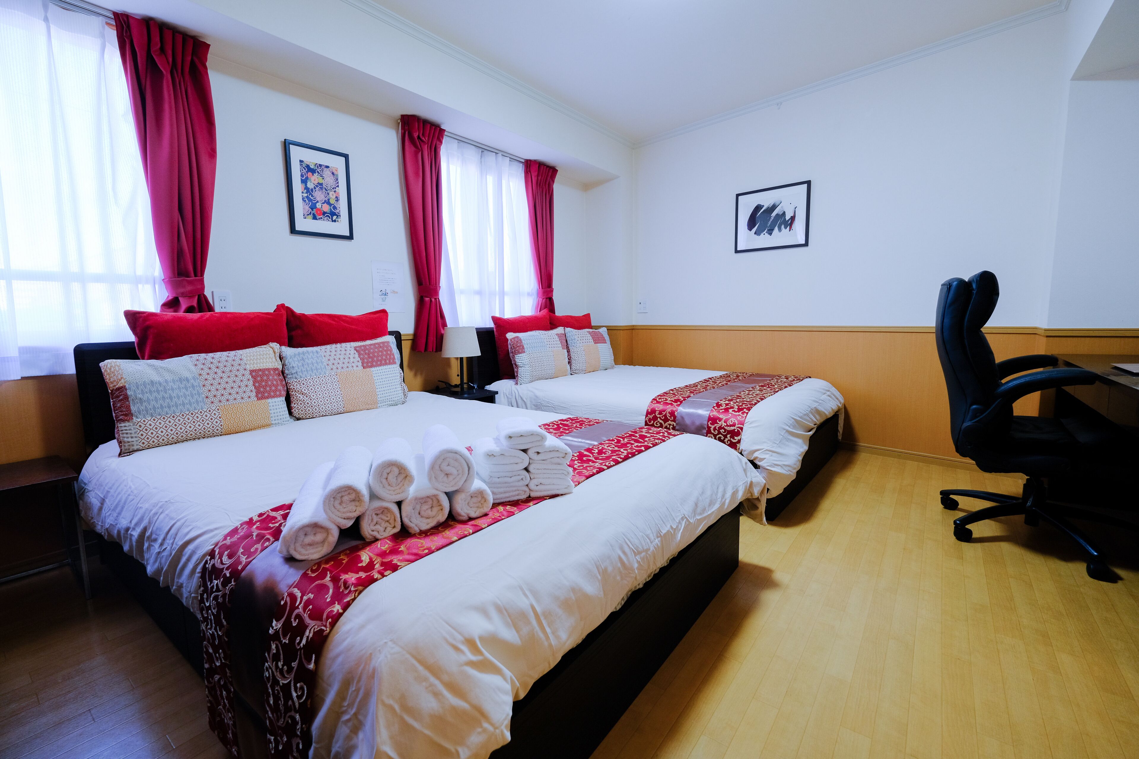 A hotel room with two beds, red and white bedding, a desk, and a chair.