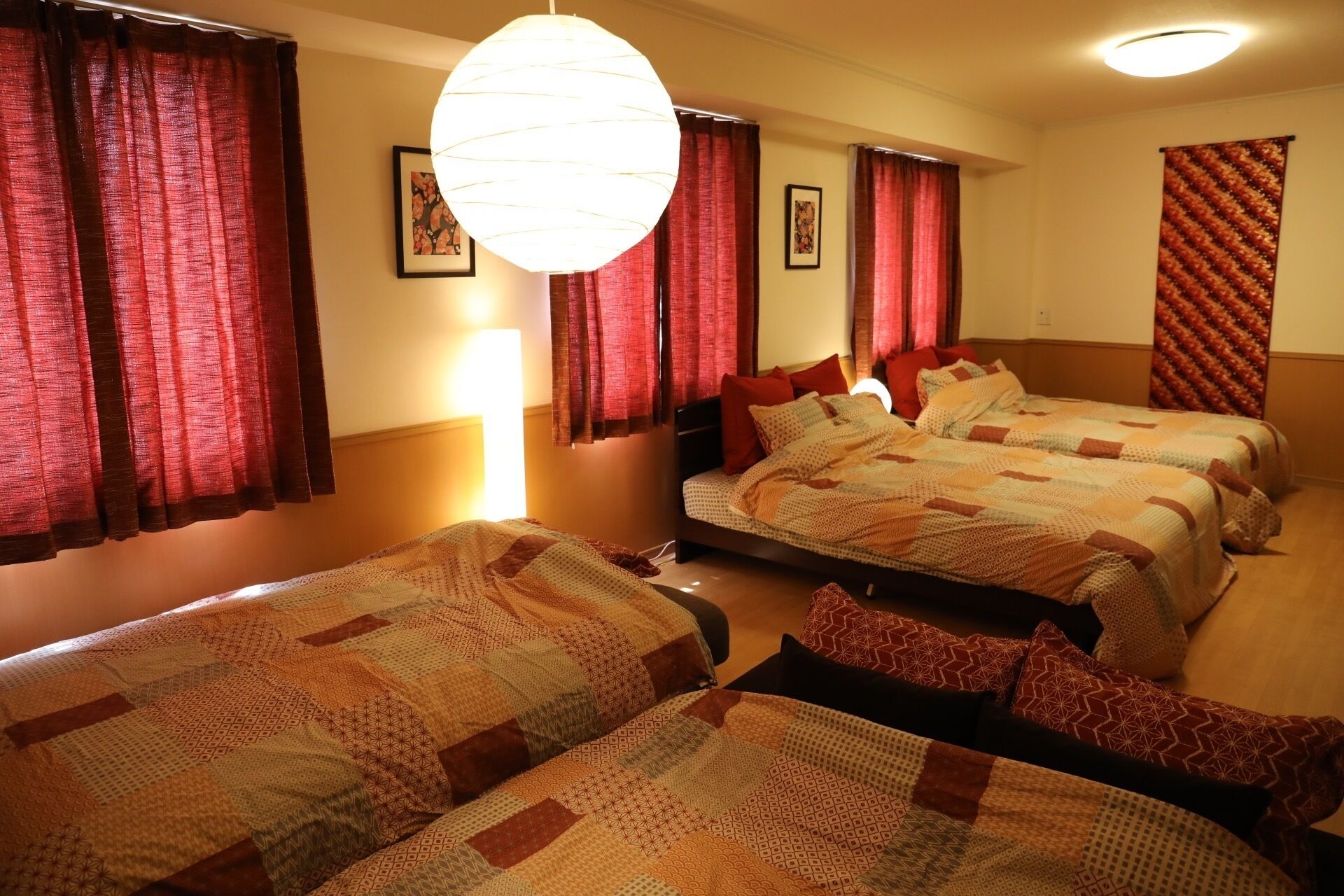 A hotel room with two beds, red curtains, a large hanging light fixture, and a patterned wall hanging.