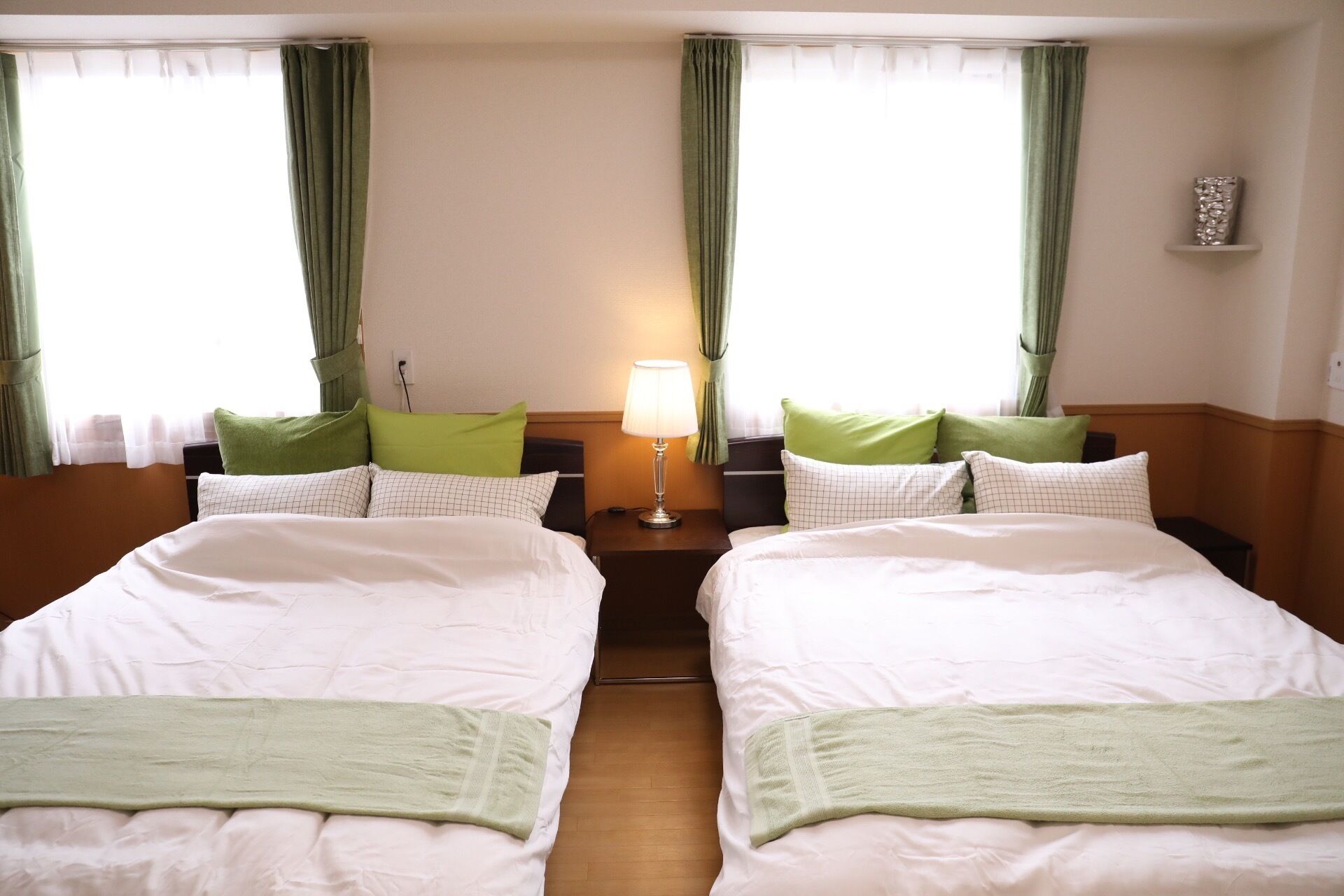 Habitación de hotel con dos camas, cada una con un almohadón verde y una manta blanca. Entre las camas, hay una mesita de noche con una lámpara.