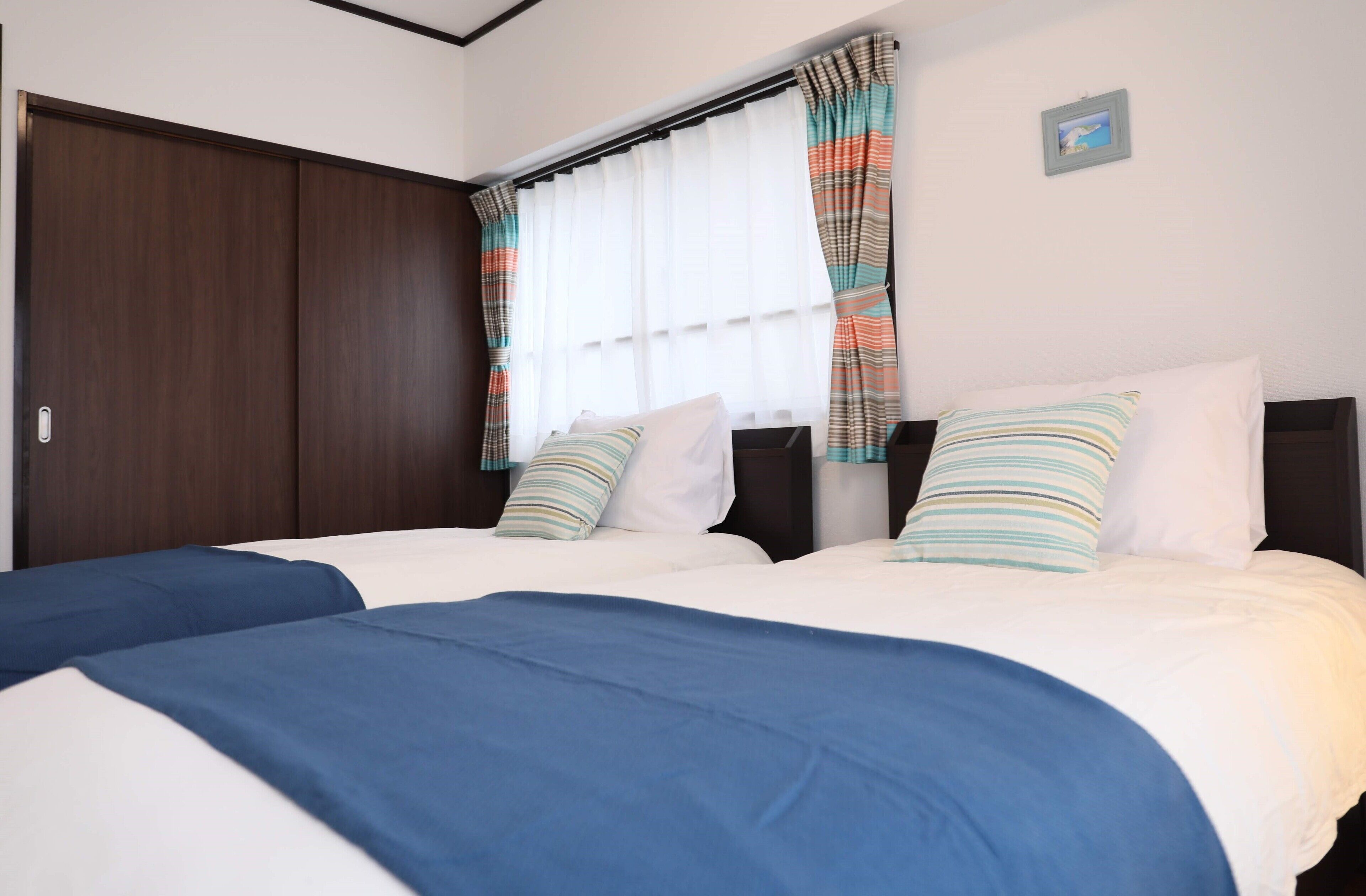 A double bed with white and blue bedding, striped pillows, and a window with white curtains and colorful drapes.
