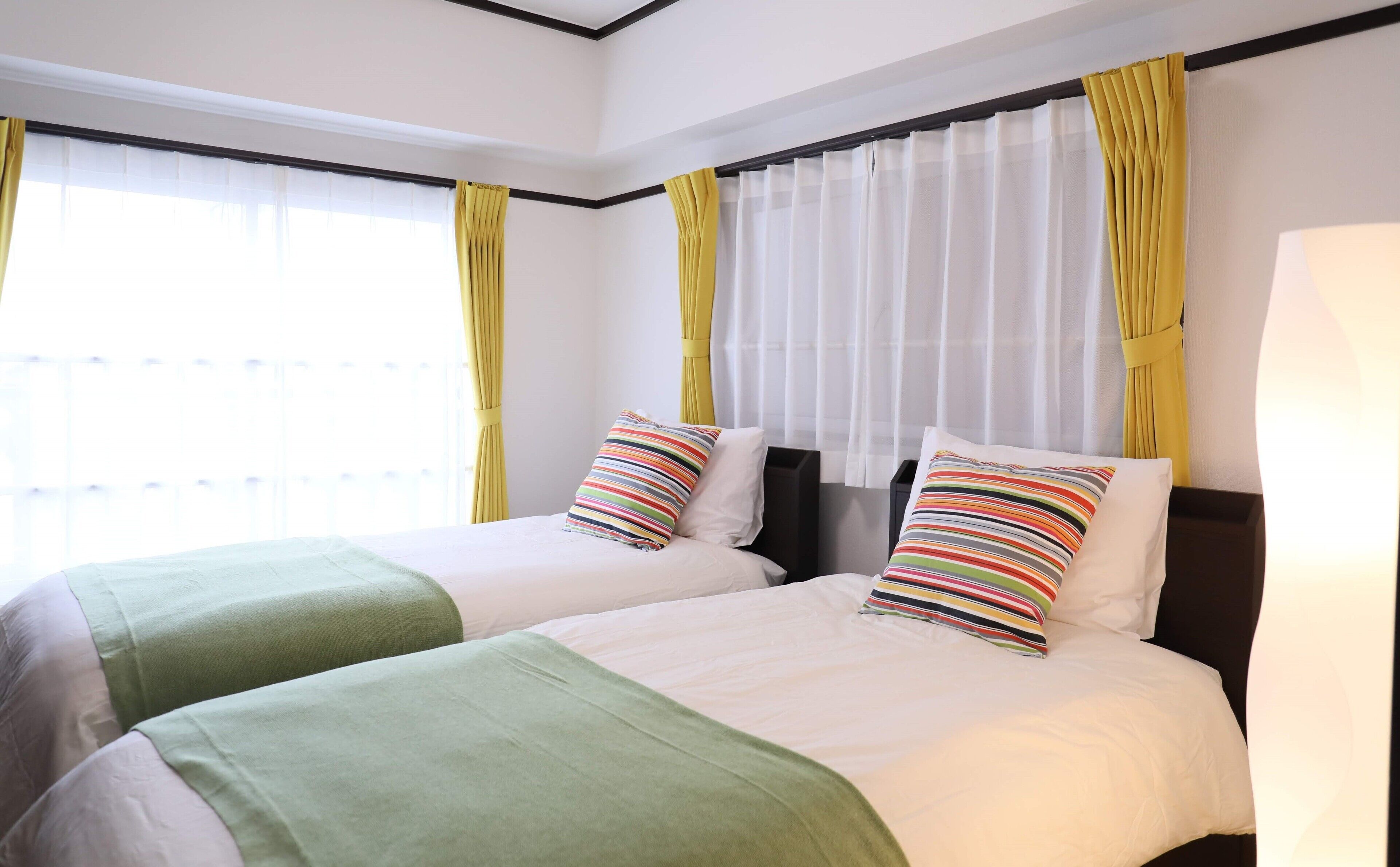 Habitación de hotel con dos camas, cada una con colcha verde y blanca y almohadas a rayas. La habitación tiene ventanas grandes con cortinas amarillas y cabecero negro.