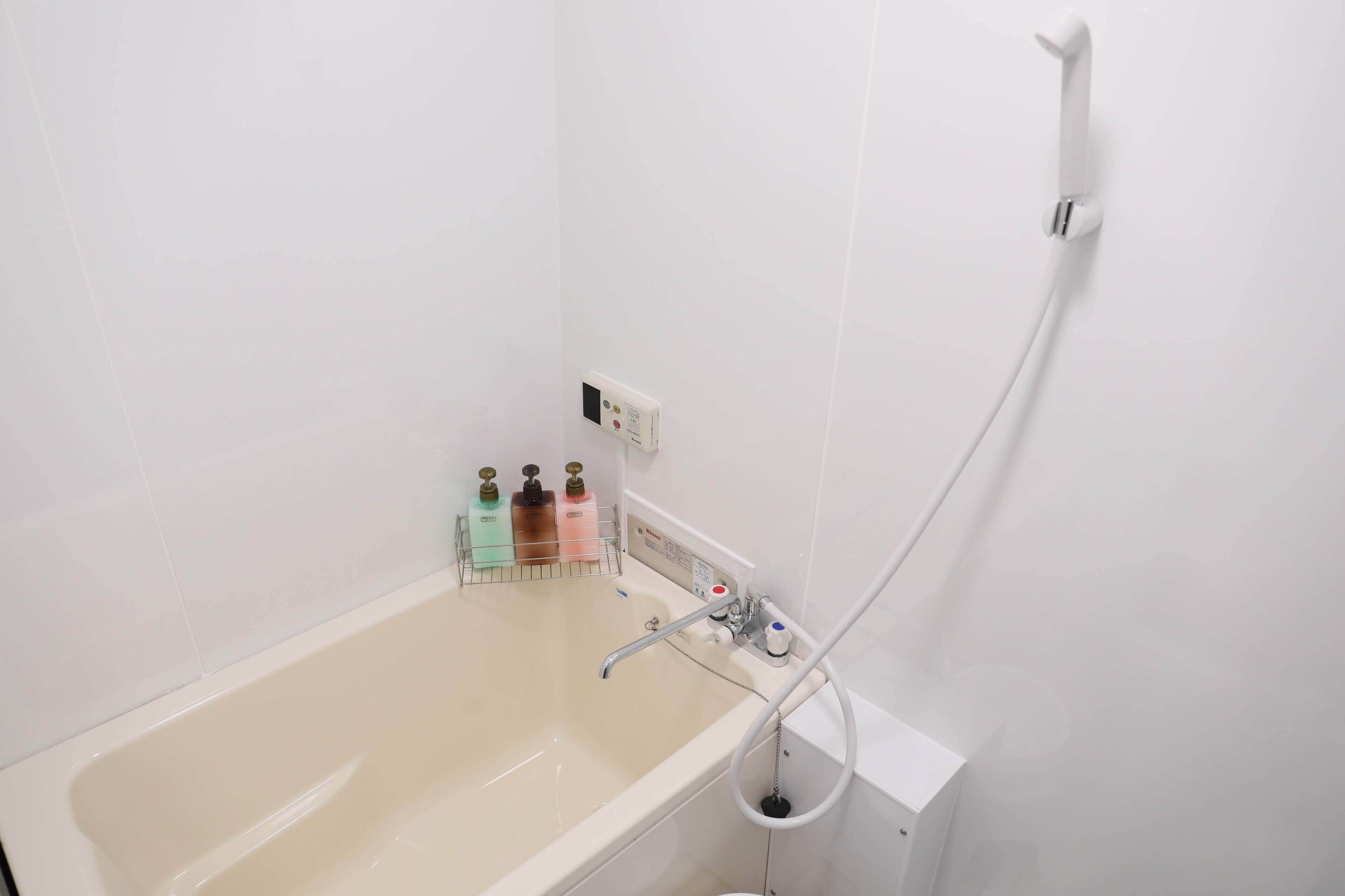 A bathroom with a bathtub, a shower, and a hand dryer. There are three bottles of soap on the wall.