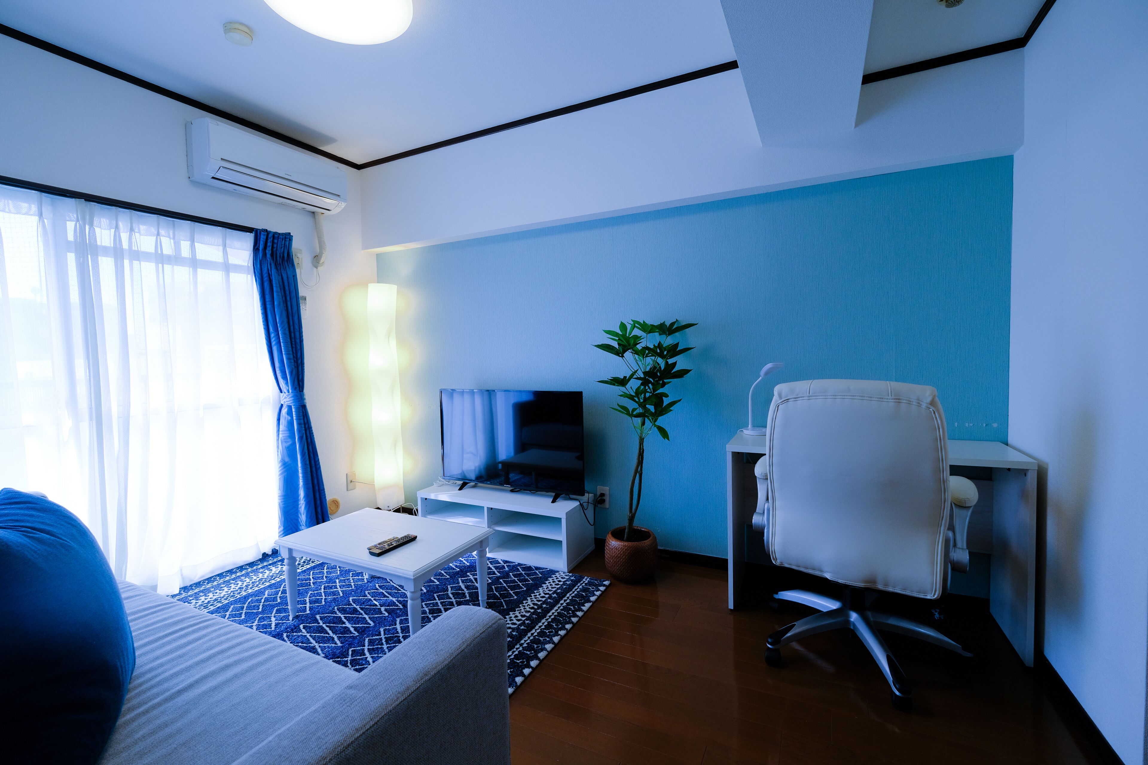 Une chambre d’hôtel moderne avec un canapé, une petite table, un téléviseur posé sur un meuble, un bureau avec une chaise et une plante en pot.