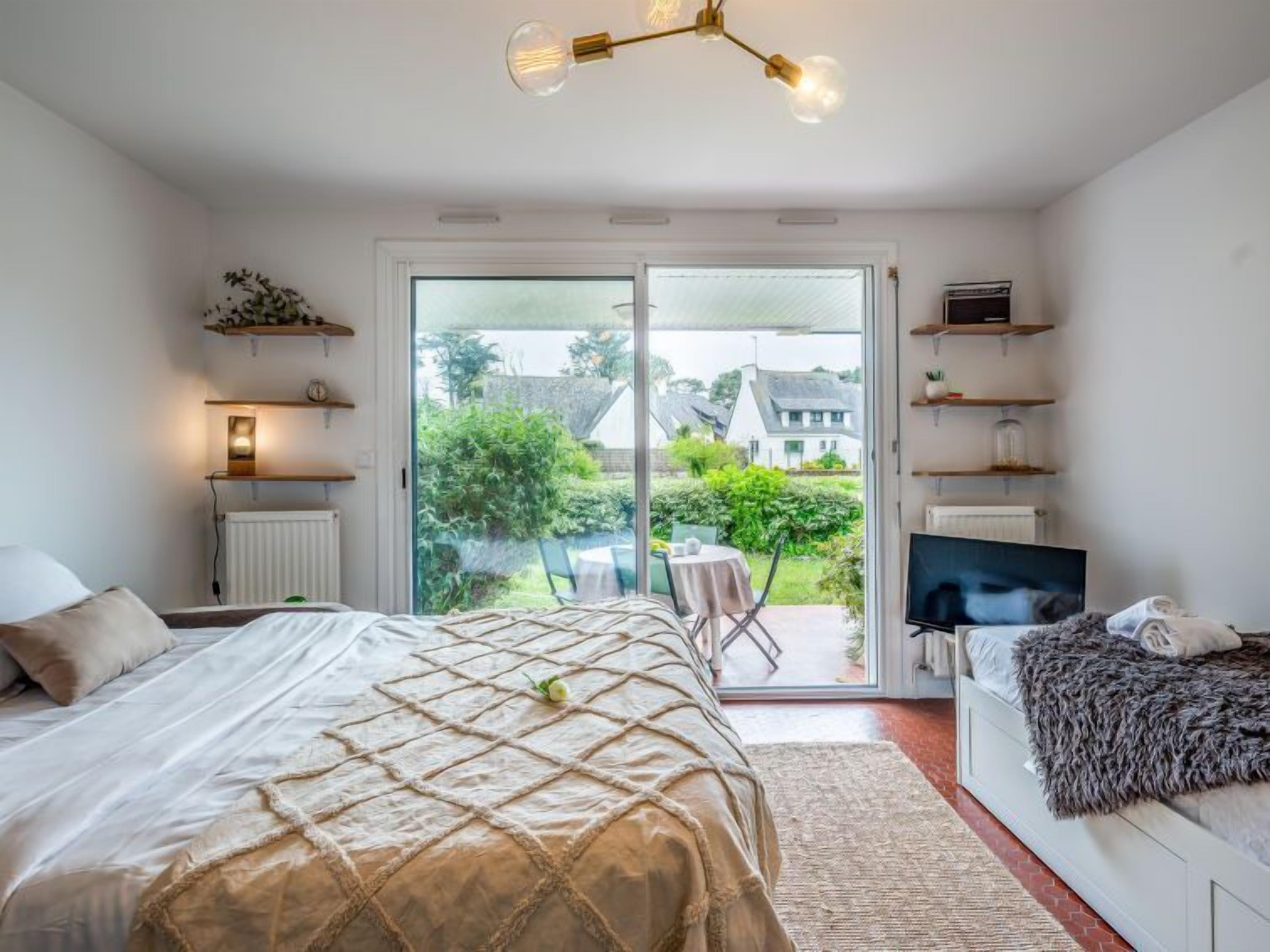 A bedroom with a bed, a TV, and a balcony with a view of houses and greenery.