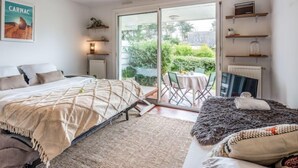 A bedroom with a bed, a sofa, a dining table with chairs, a radiator, a ceiling light fixture, a window with a view of greenery, and a framed picture on the wall. - Village de Légenèse by Interhome (Carnac)