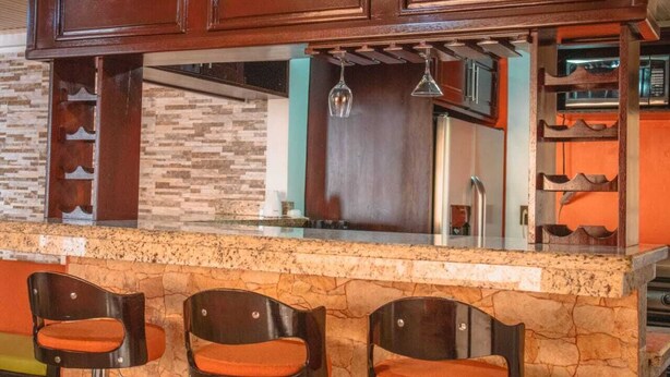 A kitchen with a stone countertop, wooden cabinets, and three bar stools with orange cushions. - The Amazing Home in a Small Town (Provincia de Alajuela)