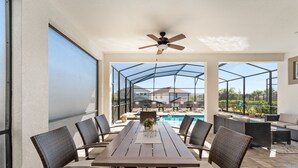 Un espacio de patio techado con una mesa y sillas, un ventilador de techo y vistas a una piscina y casas al fondo.