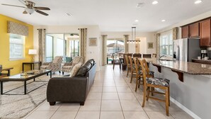 Un salón amplio con sofá, sillas y una mesa de centro. Cuenta con una zona de cocina con refrigerador y una mesa de comedor.