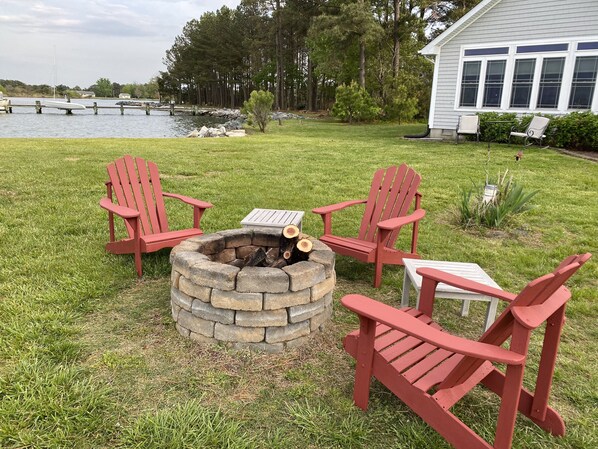 Un foyer extérieur entouré de chaises Adirondack rouges et d’une table, le tout installé sur une pelouse, avec un lac et des arbres en arrière-plan.
