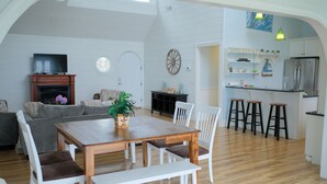 Un vaste espace de vie comprenant une table à manger en bois, des chaises blanches, une cheminée et un coin cuisine avec des tabourets.